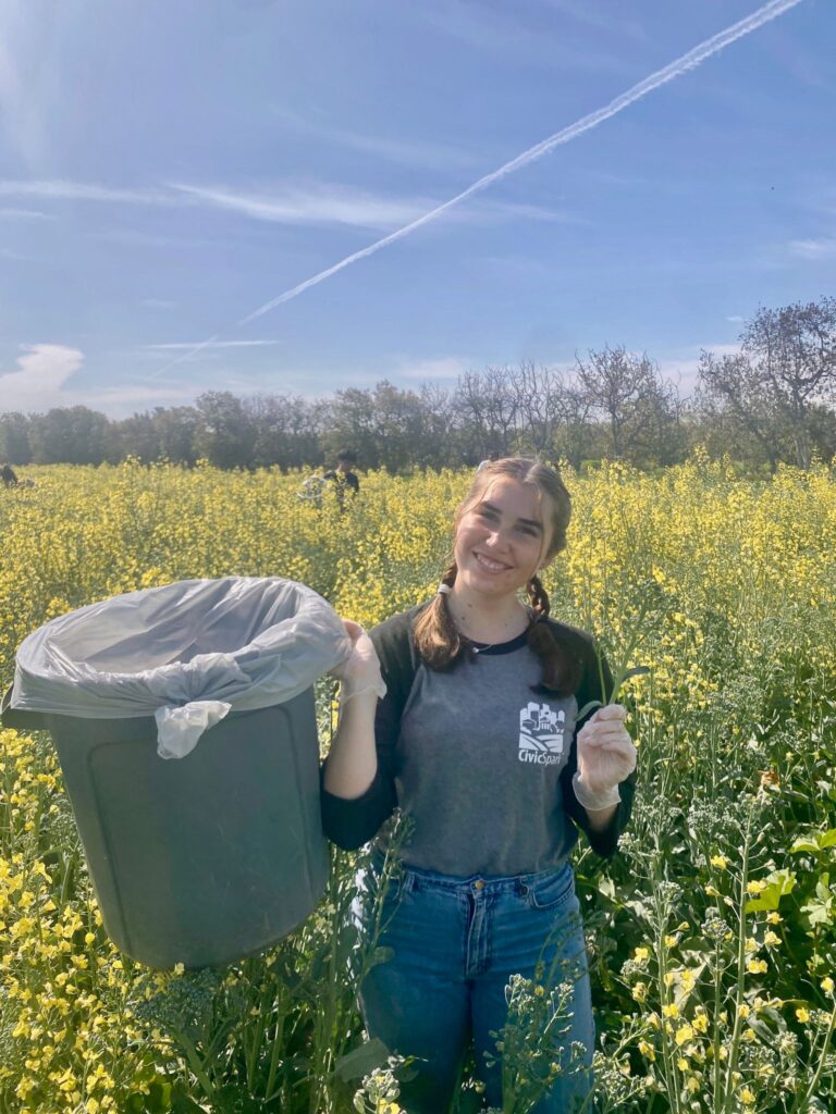 a young woman stands in a field of flowers with a trash can, picking up waste